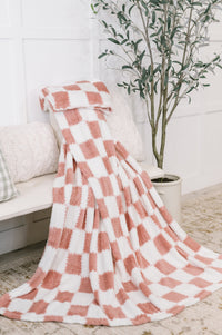 Pink and white checkered blanket draped over a chair in a cozy room with a plant and bench.