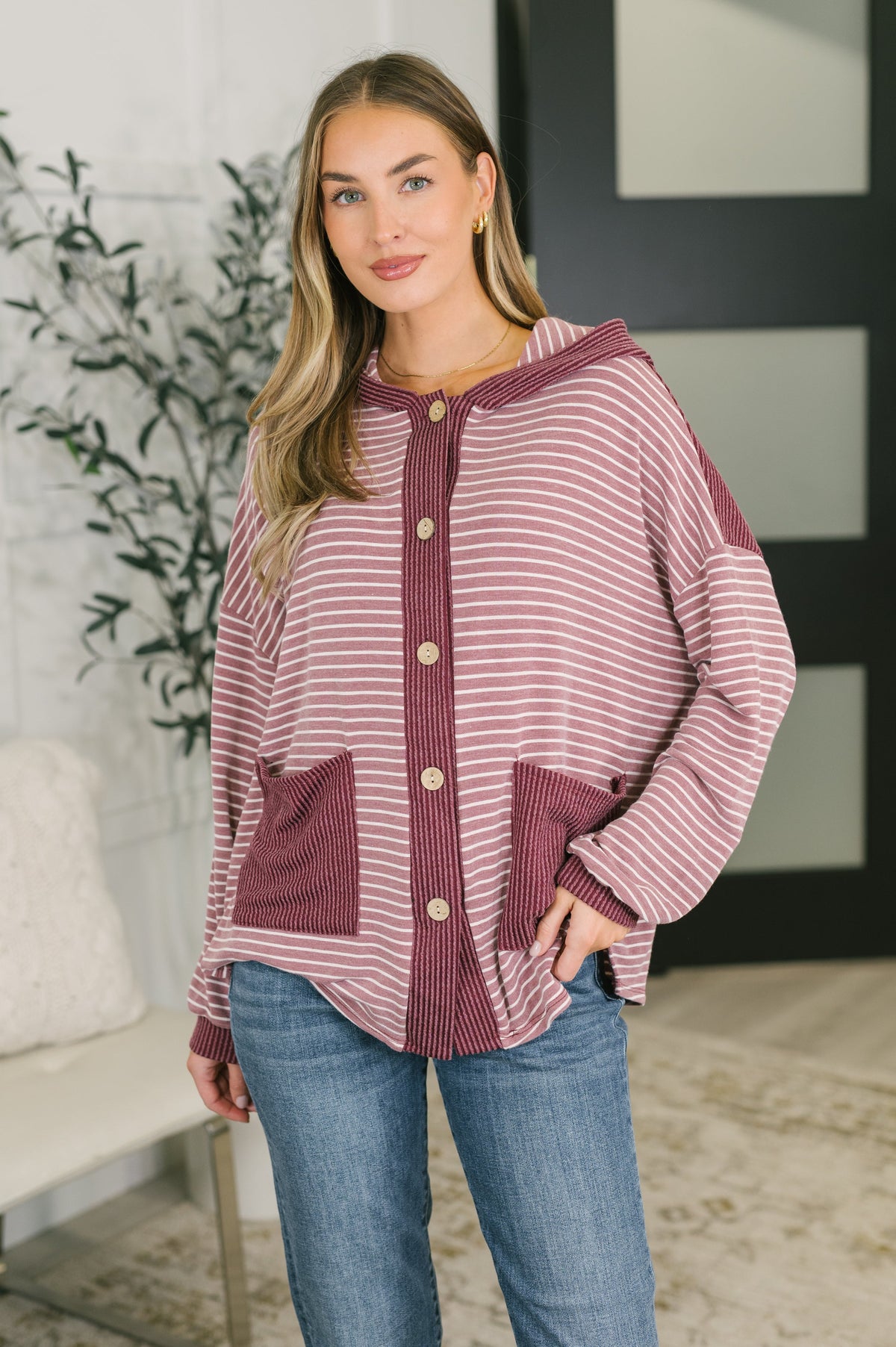 A woman wearing a dusty rose and white striped hooded cardigan with button-front closure and large front pockets, paired with blue jeans in an indoor setting.
