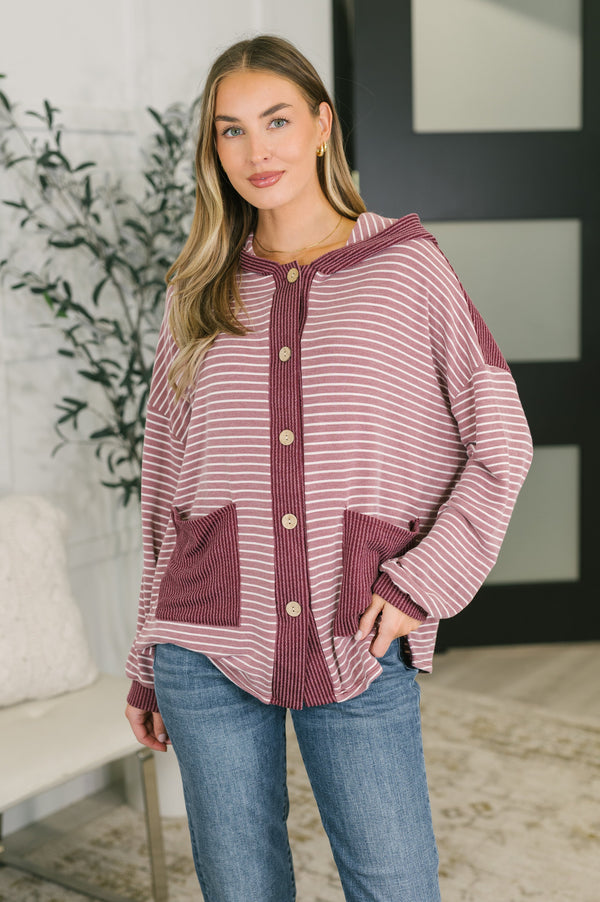 A woman wearing a dusty rose and white striped hooded cardigan with button-front closure and large front pockets, paired with blue jeans in an indoor setting.