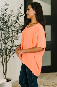 Woman wearing a peach-colored top standing indoors with a plant and wall decor in the background