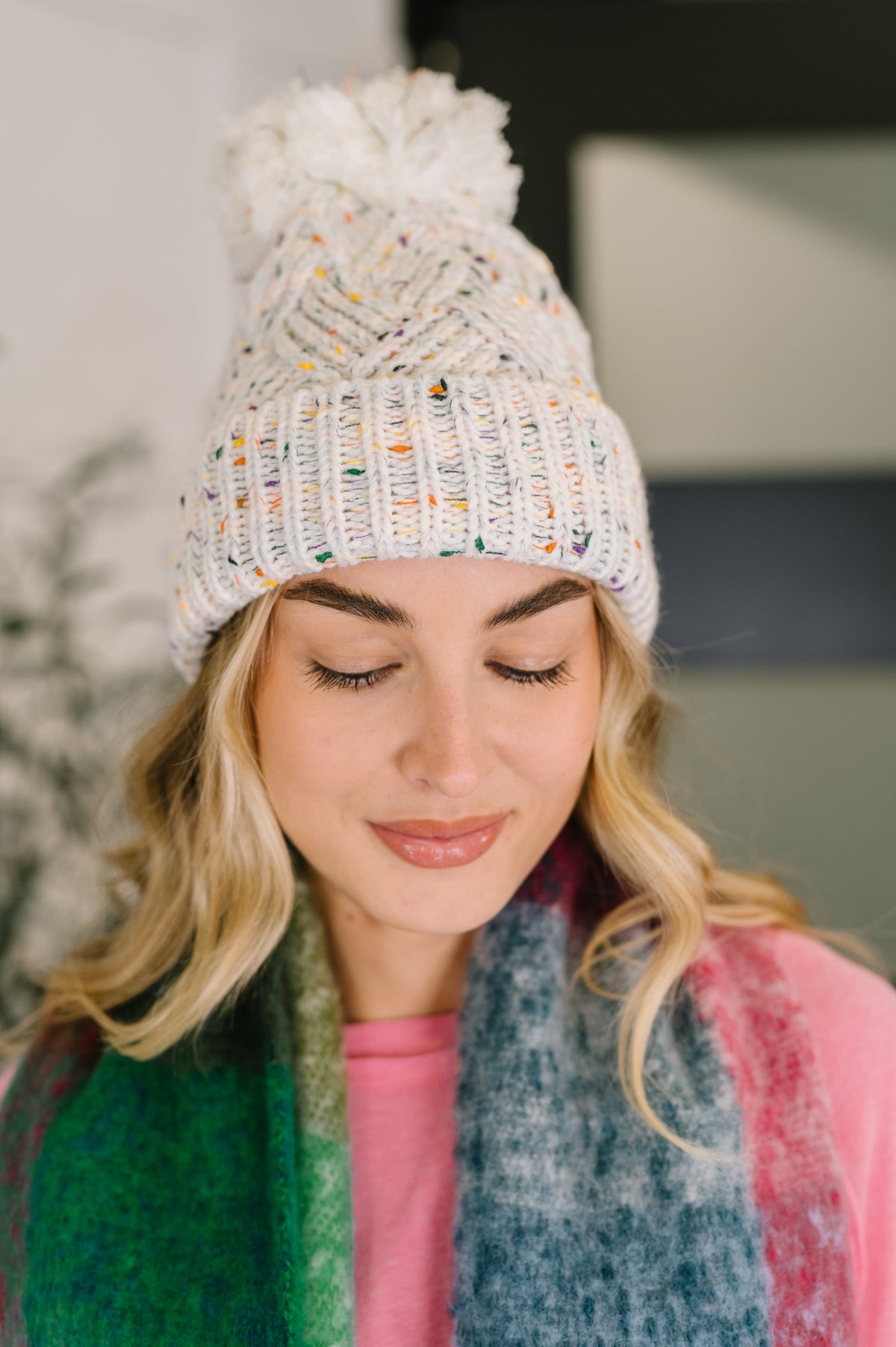 Woman wearing a knitted beanie with a pom-pom and colorful scarf, looking down.