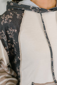 Close-up of a hooded sweatshirt with a two-tone design: one side is dark gray with a delicate floral print, and the other side is a solid cream color. The drawstrings and hem trim are also dark gray with the floral pattern.