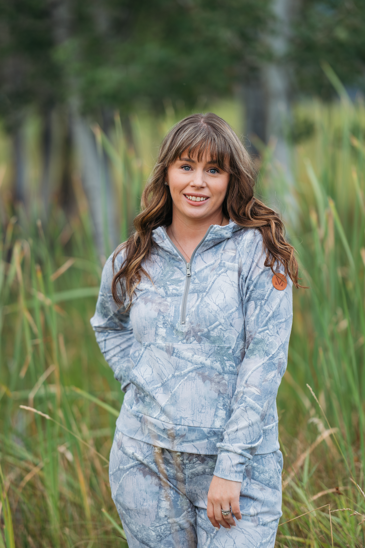 A woman standing outdoors in a grassy field wearing a light blue and white camouflage quarter-zip hoodie and matching pants.