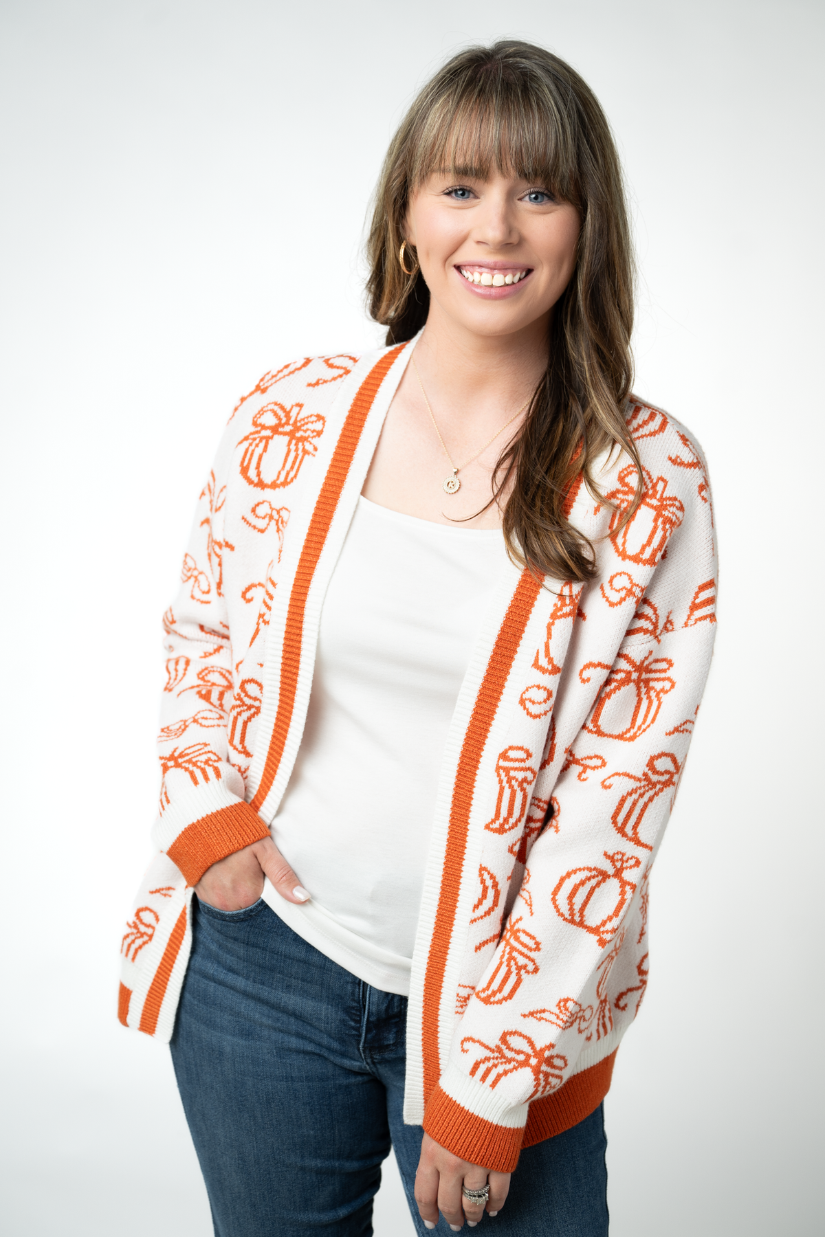 A smiling woman wearing a white knit cardigan with orange pumpkin and bow patterns, styled over a white top and blue jeans against a light gray studio background.