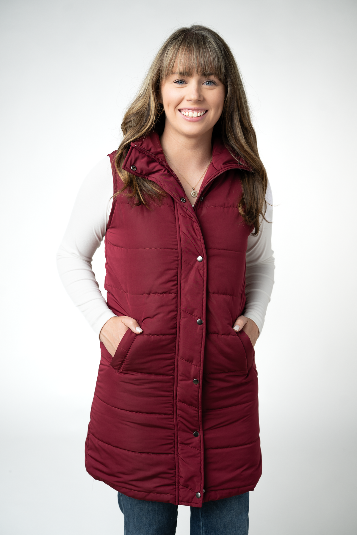 A woman wearing a sleeveless, quilted burgundy puffer vest over a white long-sleeve shirt and blue jeans, standing against a light gray studio background.