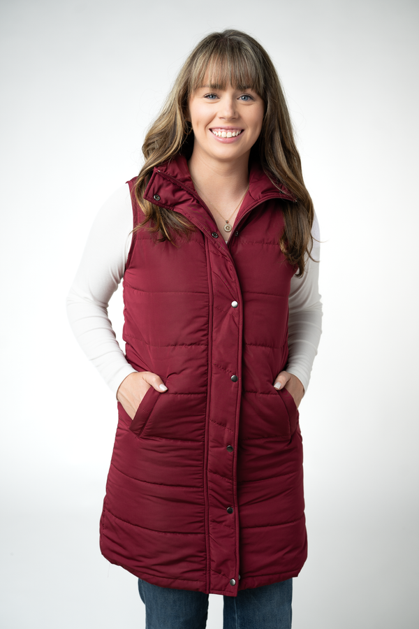 A woman wearing a sleeveless, quilted burgundy puffer vest over a white long-sleeve shirt and blue jeans, standing against a light gray studio background.