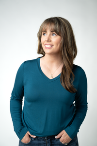 A woman with brown hair and bangs wearing a teal long-sleeve V-neck top, standing against a light gray studio background with her hands in her jeans pockets.
