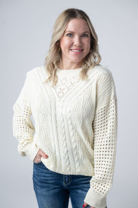 Woman wearing a cream cable knit sweater and blue jeans against a plain background