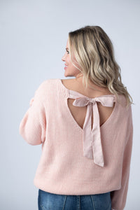 Woman wearing a pink sweater with a bow detail on the back against a light gray background