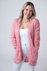 Woman wearing a pink cable knit cardigan over a white top and pants on a light gray background