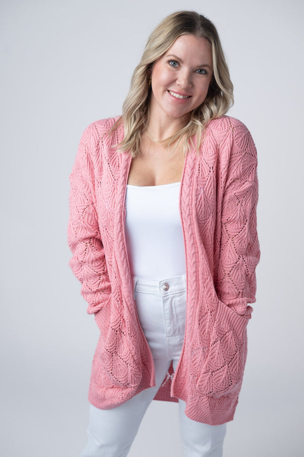 Woman wearing a pink cable knit cardigan over a white top and pants on a light gray background