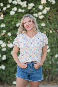 Woman wearing a floral top and denim shorts standing in front of a flowering bush.
