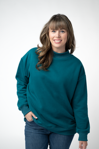 A smiling woman with brown hair and bangs wearing an oversized teal crewneck sweatshirt and blue jeans, standing against a light gray studio background.