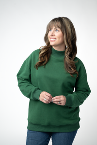 A woman with long brown hair wearing a dark green mock neck sweatshirt and blue jeans, standing against a light gray studio background.