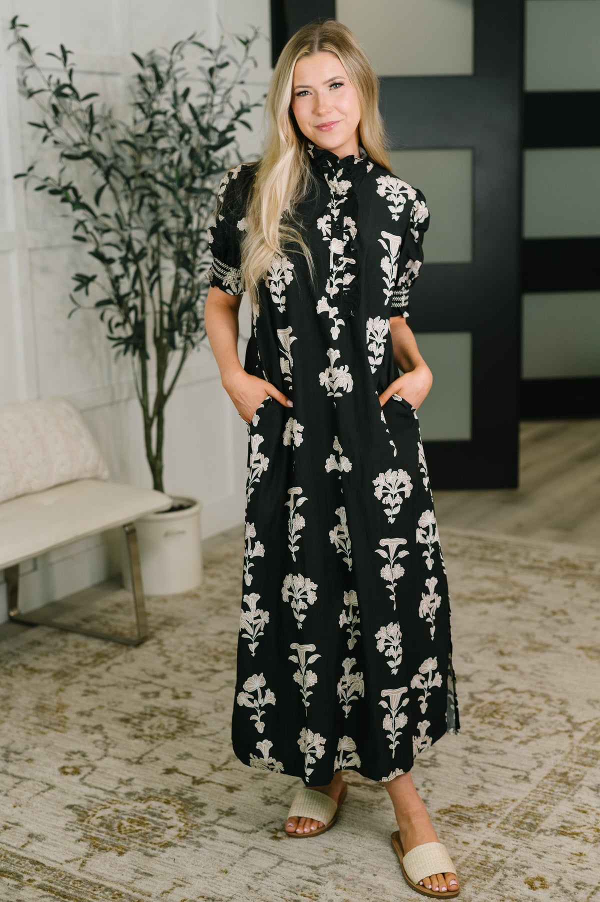 A woman modeling a black midi dress with a white floral pattern, standing in a styled indoor room with a bench, potted plant, and patterned rug.