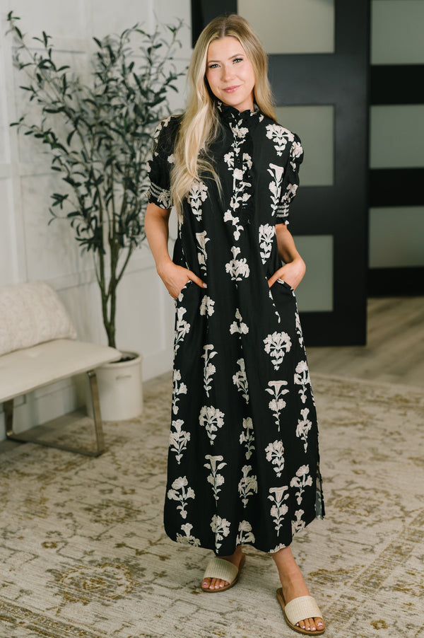 A woman modeling a black midi dress with a white floral pattern, standing in a styled indoor room with a bench, potted plant, and patterned rug.