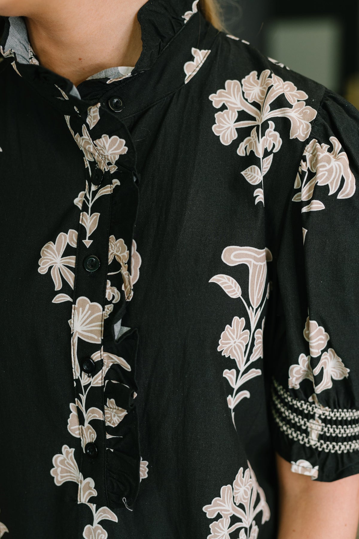 Close-up of a black short-sleeve blouse with a beige floral print, featuring a high collar, button placket, and patterned sleeve cuff.