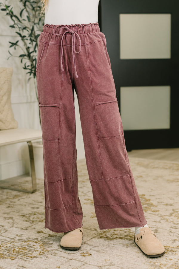 A person wearing dusty rose wide-leg drawstring pants with patchwork paneling and beige slip-on shoes, standing on a patterned rug in an indoor room setting.