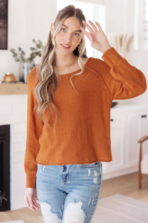 A woman wearing a fuzzy long-sleeve crewneck sweater in burnt orange, paired with distressed light blue jeans, standing in a bright, modern living room.