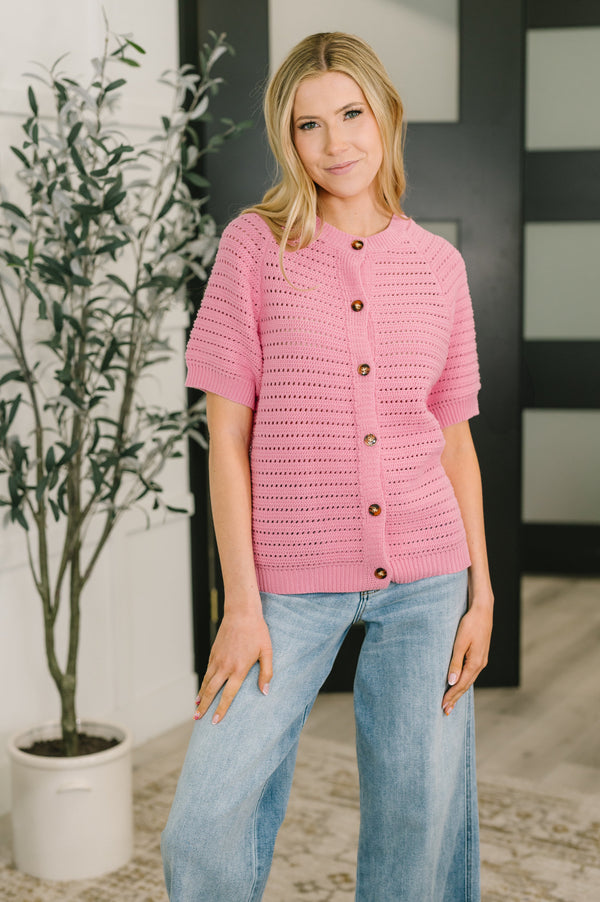 Woman wearing a pink crochet cardigan with buttons, standing indoors next to a plant.