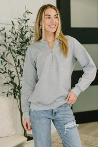 Woman wearing a gray sweater and light blue jeans standing indoors with a plant in the background.