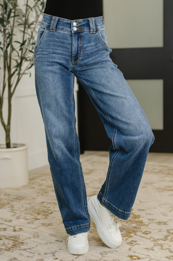 Person wearing blue jeans and white sneakers in a room with a plant and wall.