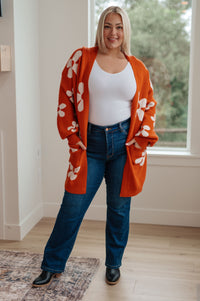 Woman wearing an orange floral cardigan, white top, and blue jeans indoors.