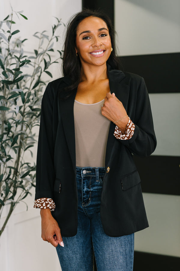 Woman wearing a black blazer, beige top, and blue jeans with a plant in the background