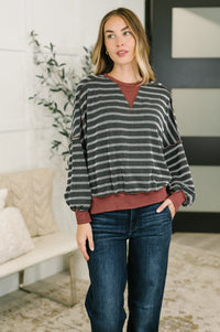 A woman modeling a black and white striped long-sleeve top with a rust-colored v-neck and hem, paired with dark blue jeans in a modern indoor setting.