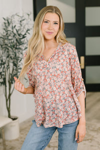 A woman with long blonde hair wearing a loose, three-quarter sleeve floral blouse and blue jeans, standing in a brightly lit indoor setting with a potted plant and a modern black door in the background.