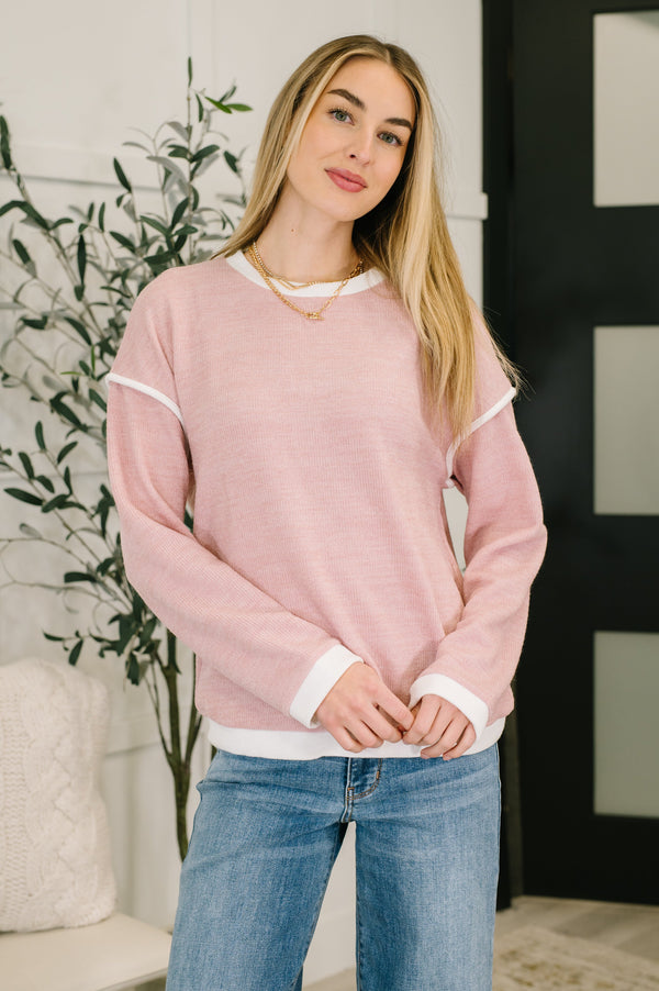 Woman wearing a pink sweater and blue jeans standing indoors with a plant in the background