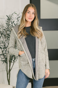 A woman modeling a light gray and white textured open-front cardigan with a solid gray hood, worn over a dark gray top and blue jeans in an indoor setting.