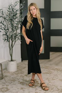 Woman wearing a black dress standing indoors with a plant and decorative wall in the background