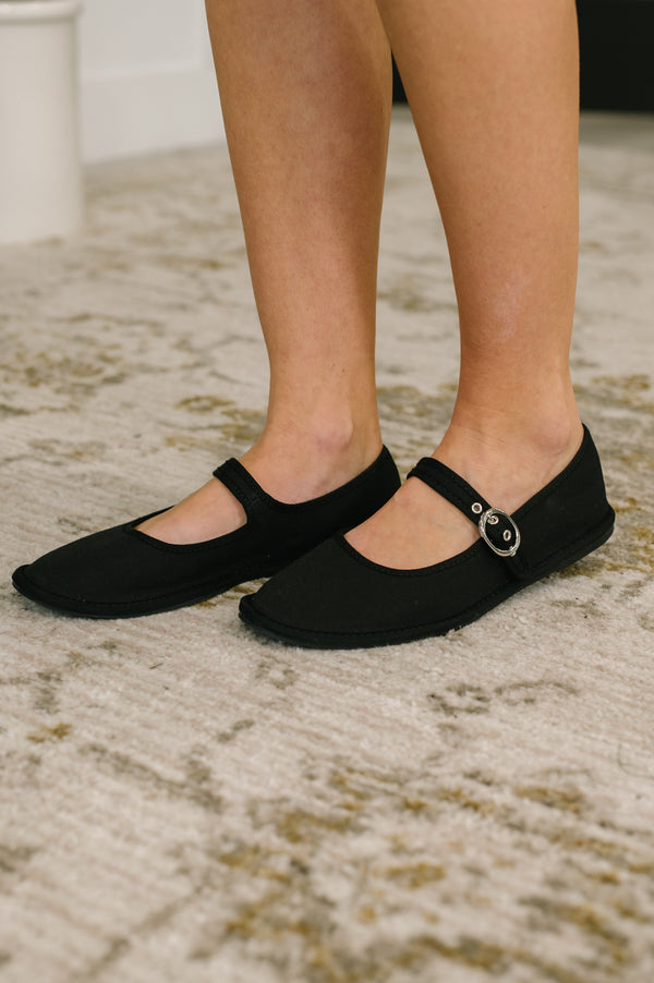 Black flat shoes worn by a person on a textured floor.