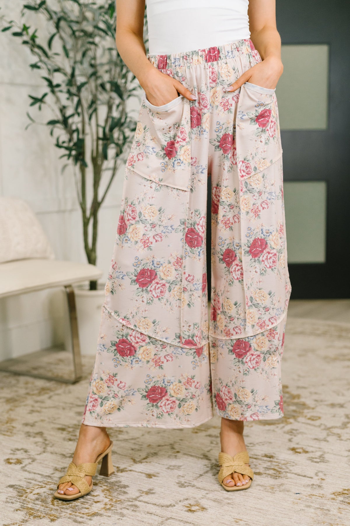 A person wearing high-waisted, wide-leg cropped pants with a pink and red floral print on a light beige background, styled with a white top and tan heeled sandals in an indoor setting.
