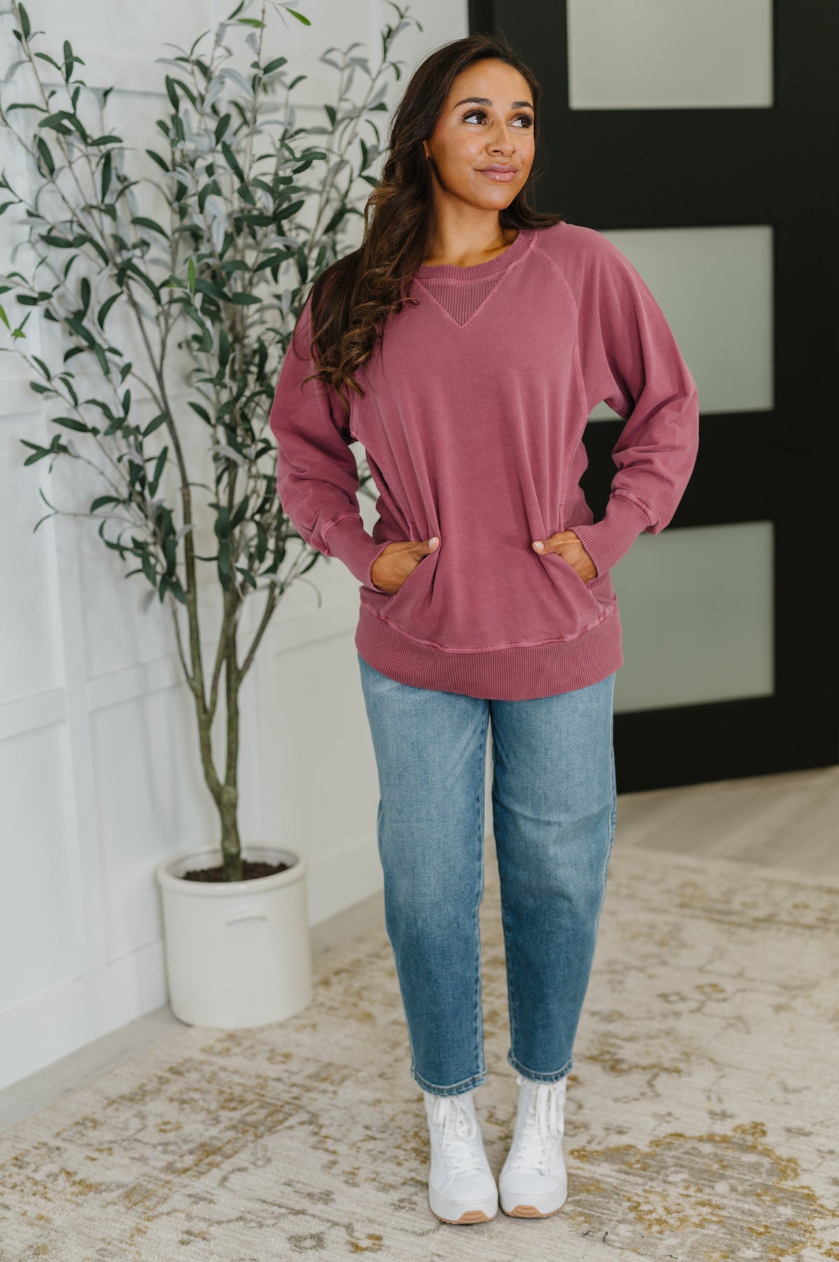 Woman wearing a pink sweatshirt and blue jeans standing indoors next to a plant.