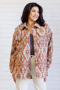 A woman modeling a patterned button-up jacket with a rust orange and cream geometric design, worn over a dark top and paired with beige pants, standing against a white brick wall.