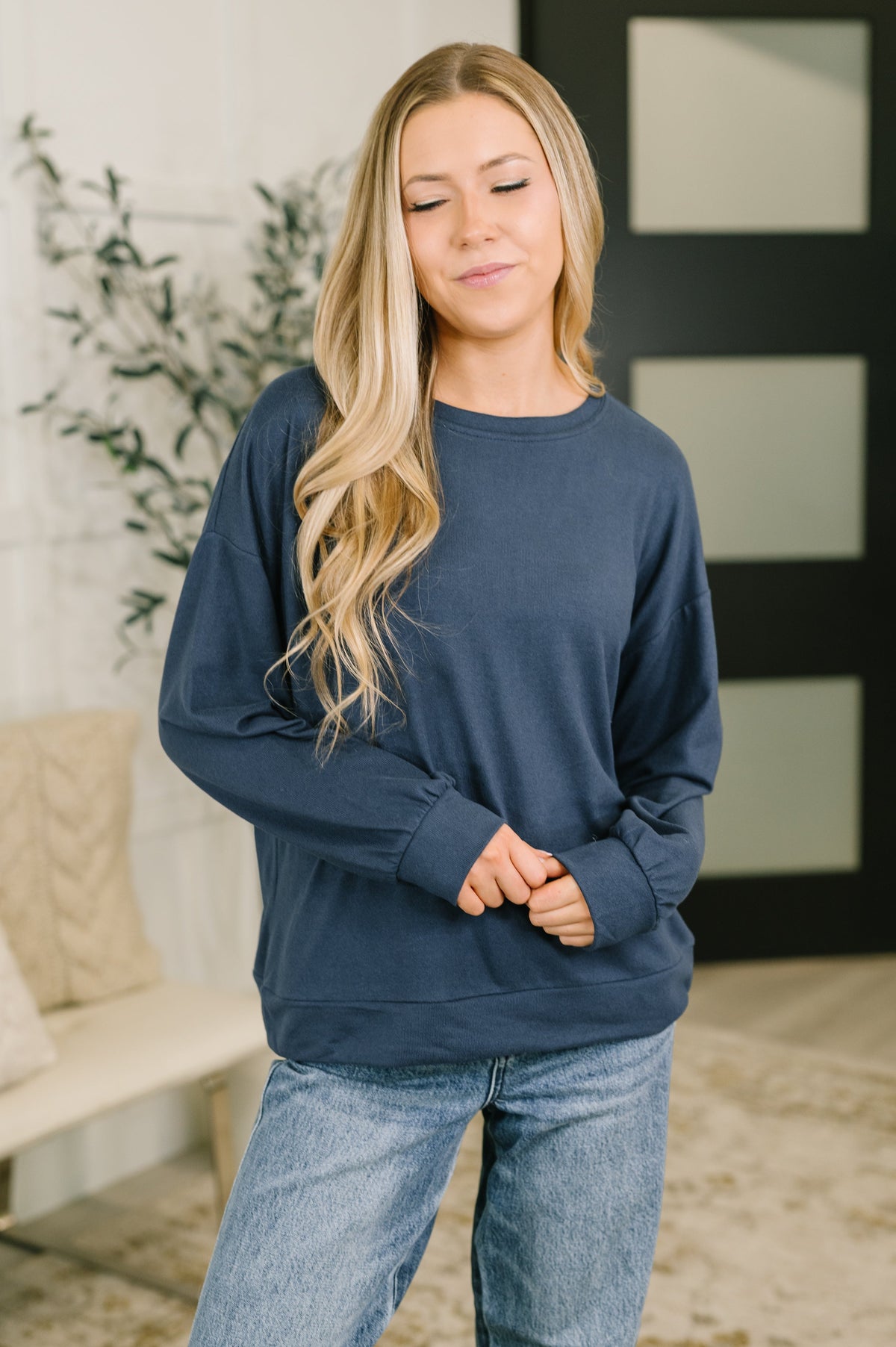 A woman with long blonde hair wearing a navy blue crewneck sweatshirt and light blue jeans, standing in a brightly lit indoor setting.