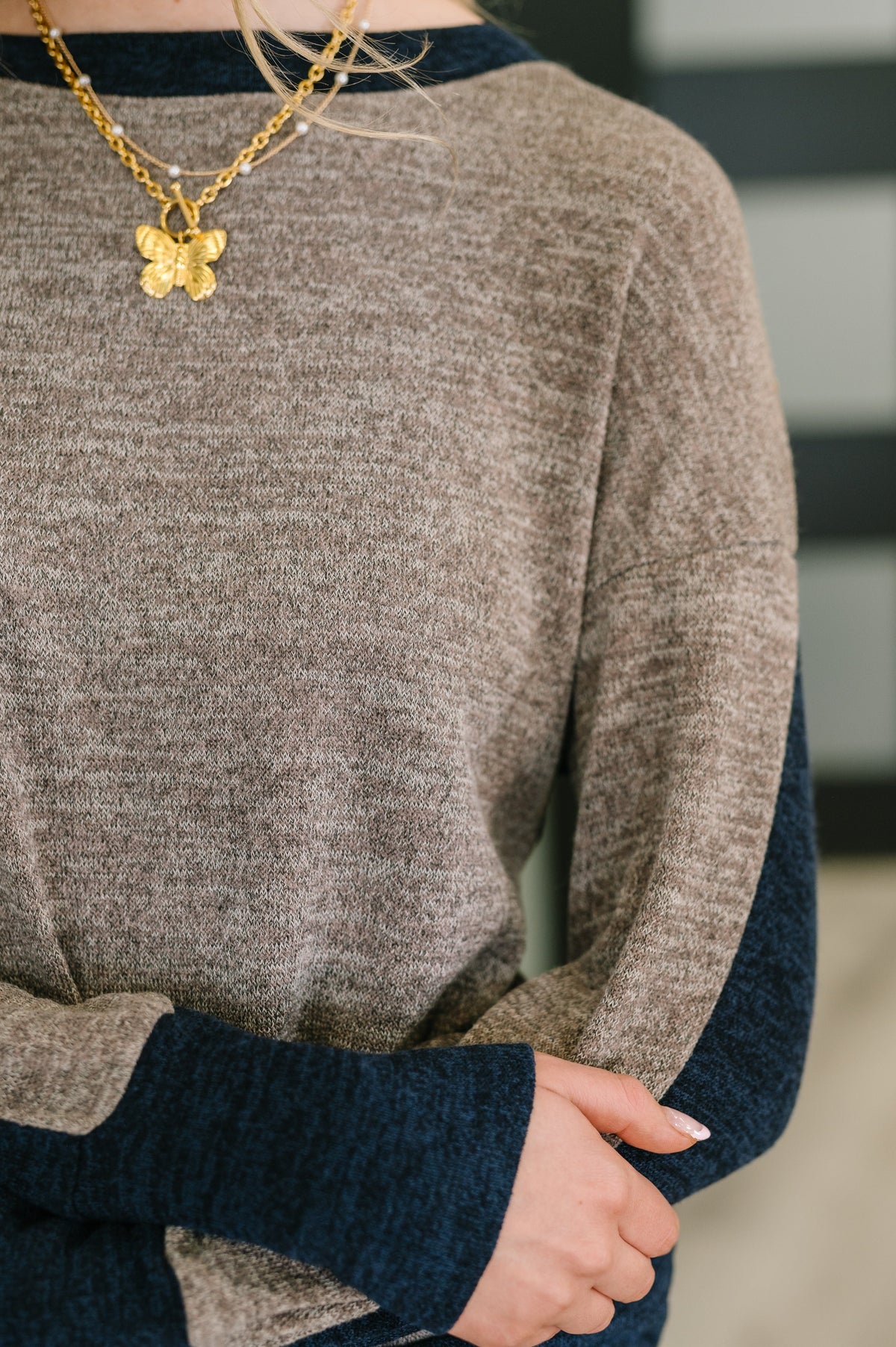 Close-up of a person wearing a gold necklace with a butterfly pendant, brown sweater, and blue sleeves.