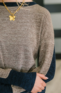 Close-up of a person wearing a gold necklace with a butterfly pendant, brown sweater, and blue sleeves.