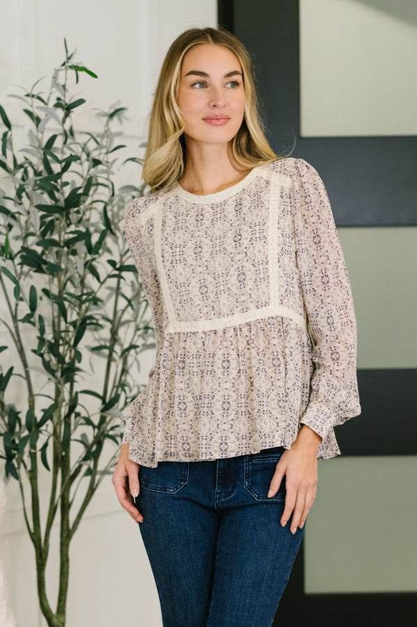 Woman wearing a patterned blouse and jeans standing indoors with a plant in the background