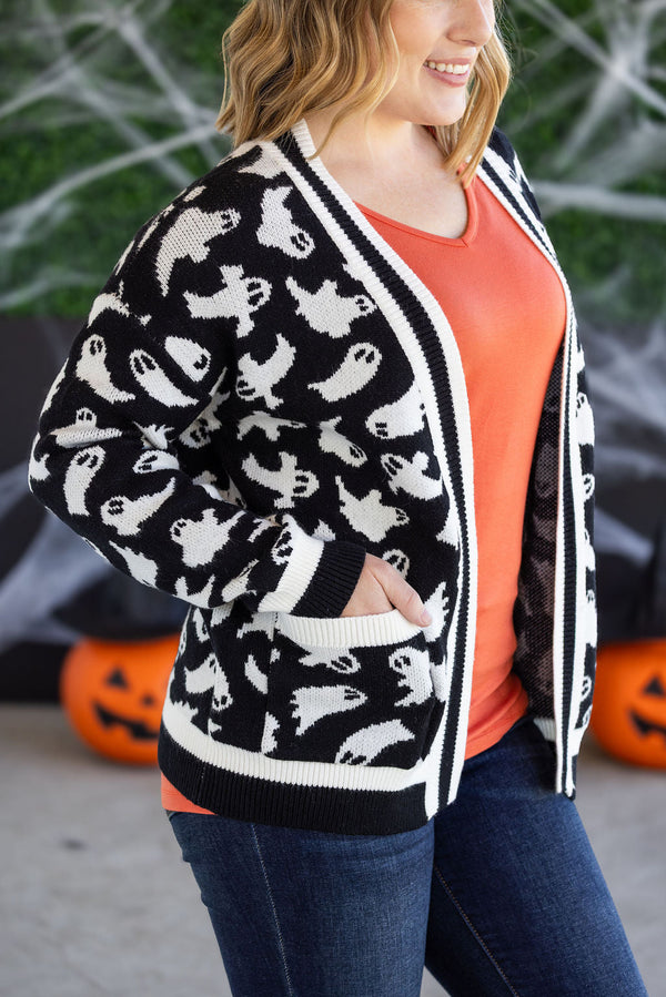 Person wearing a black cardigan with ghost pattern in front of Halloween decorations.