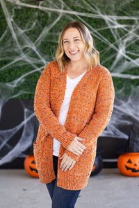 Woman wearing an orange cardigan in front of a Halloween-themed background with pumpkins and spider webs.