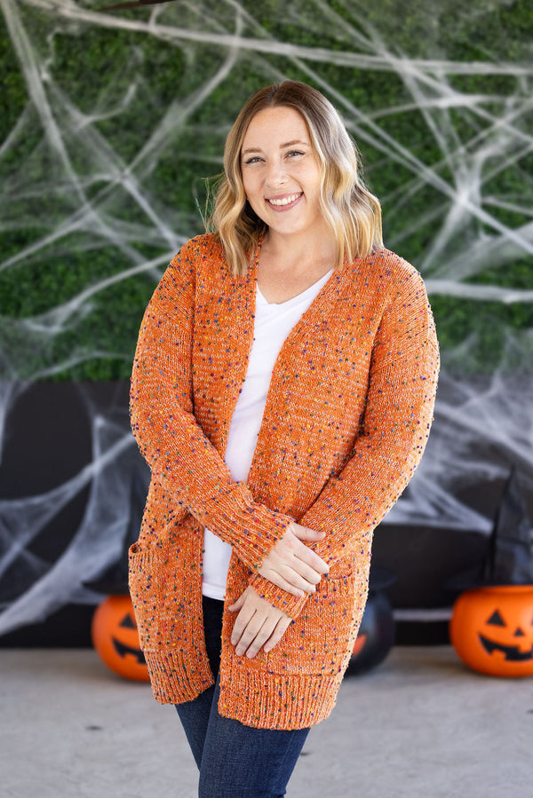 Woman wearing an orange cardigan in front of a Halloween-themed background with pumpkins and spider webs.