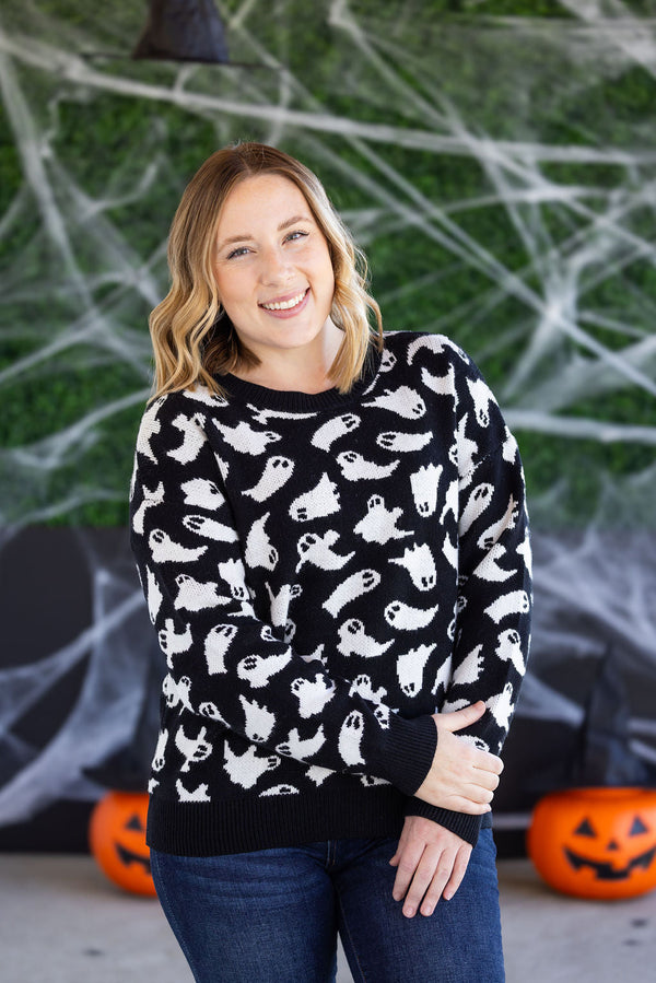 Woman wearing a black sweater with white ghost patterns in front of Halloween decorations.