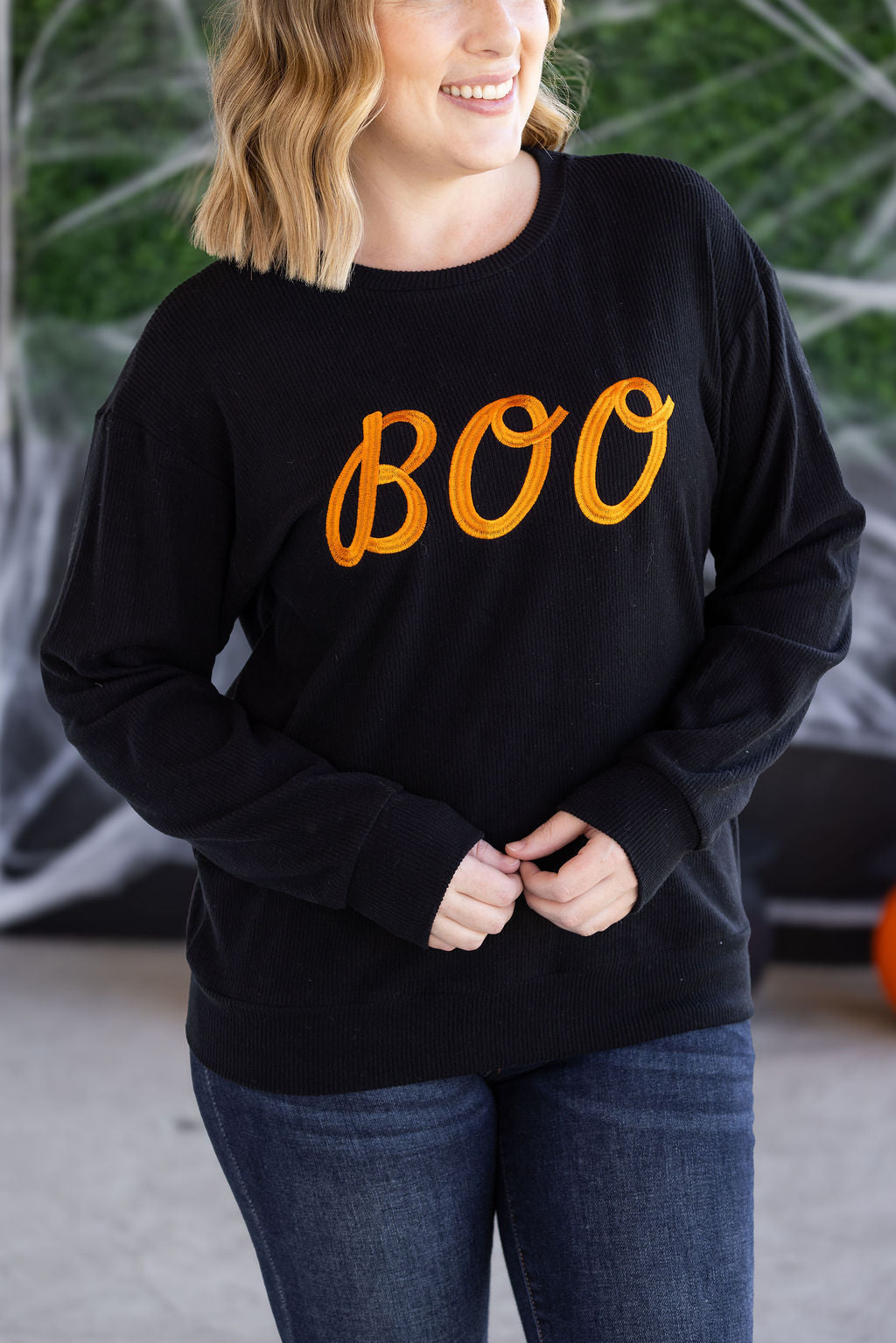 Person wearing a black sweatshirt with 'BOO' in orange text, standing against a blurred green background.