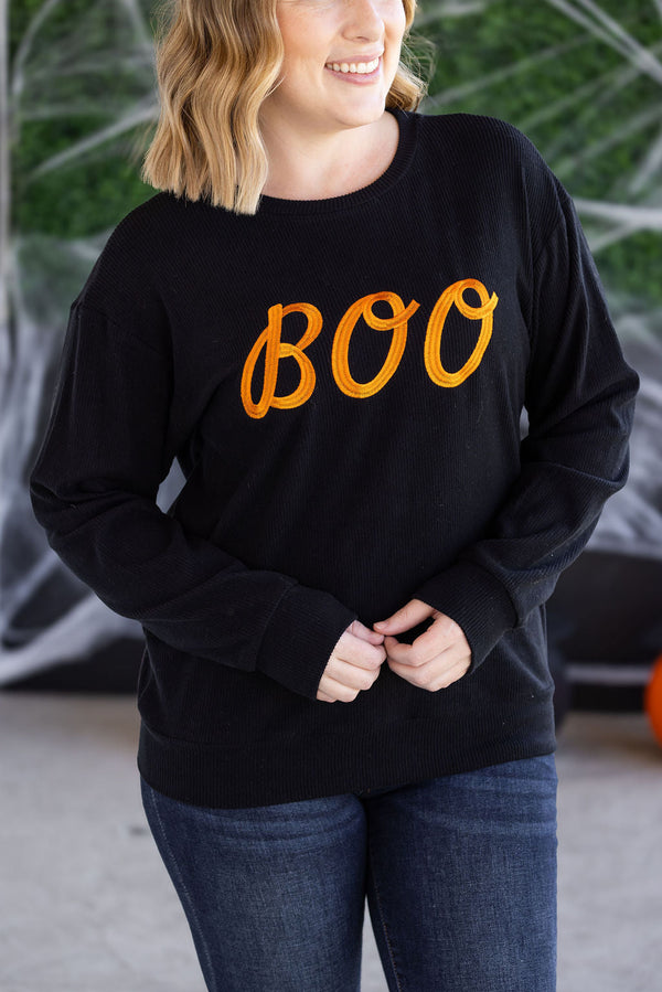 Person wearing a black sweatshirt with 'BOO' in orange text, standing against a blurred green background.