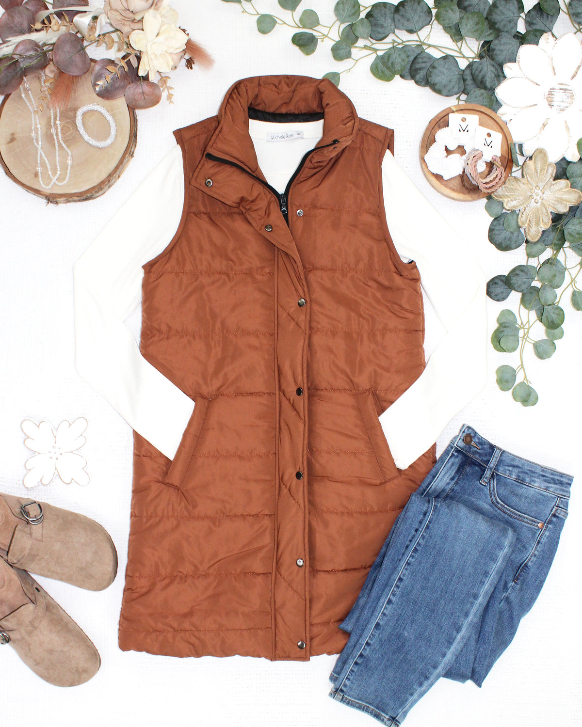 A rust-brown sleeveless puffer vest styled in a flat lay with a white long-sleeve shirt, blue jeans, brown suede shoes, and decorative floral props on a white background.