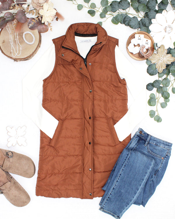 A rust-brown sleeveless puffer vest styled in a flat lay with a white long-sleeve shirt, blue jeans, brown suede shoes, and decorative floral props on a white background.