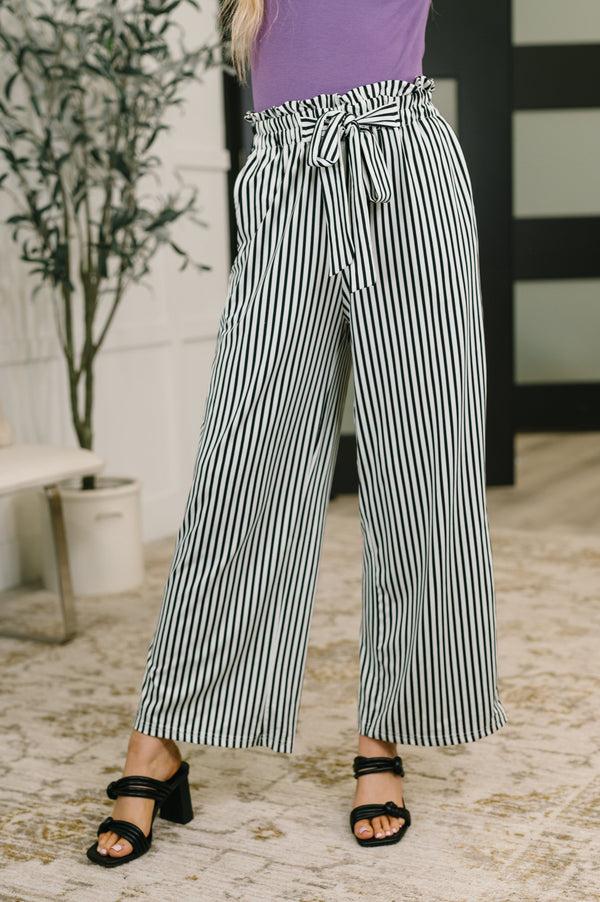 A person wearing high-waisted, wide-leg pants with vertical black and white stripes, a purple top, and black heeled sandals in an indoor setting.